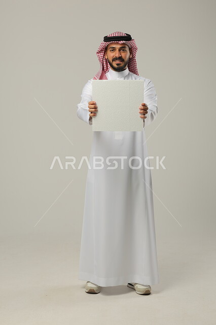 النظر إلى الكاميرا بإيماءات السعادة و السرور, استخدام لوحات توضيحية و وسائل تعليمية, بورتريه لرجل عربي خليجي سعودي يرتدي الثوب التقليدي و الشماغ يمسك بيديه لوحة بيضاء فارغة, صورة للجسم بالطول الكامل, خلفية رمادية