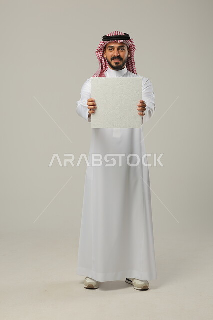 استخدام لوحات توضيحية و وسائل تعليمية, العروض والإعلانات التسويقية, بورتريه لرجل عربي خليجي سعودي يرتدي الثوب التقليدي و الشماغ يمسك بيديه لوحة بيضاء فارغة وينظر إلى الكاميرا, صورة للجسم بالطول الكامل, خلفية رمادية