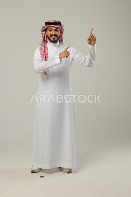 الوقوف باستقامة و النظر إلى الكاميرا بإيماءات السعادة و السرور, الإشارة إلى شيء ما, بورتريه لرجل عربي خليجي سعودي يرتدي الثوب التقليدي و الشماغ يشير بإصبعي السبابة إلى الأعلى, صورة للجسم بالطول الكامل, خلفية رمادية