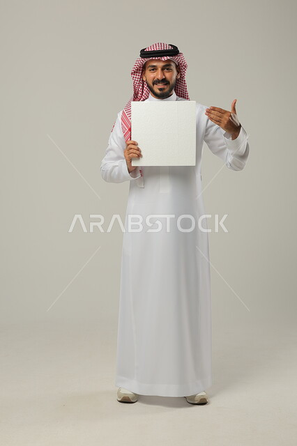 الترويج الإعلاني على موك اب أبيض, التسويق للخدمات و المنتجات, بورتريه لرجل عربي خليجي سعودي يرتدي الثوب التقليدي و الشماغ يشير بيده إلى اللوحة البيضاء الفارغة, صورة للجسم بالطول الكامل, خلفية رمادية