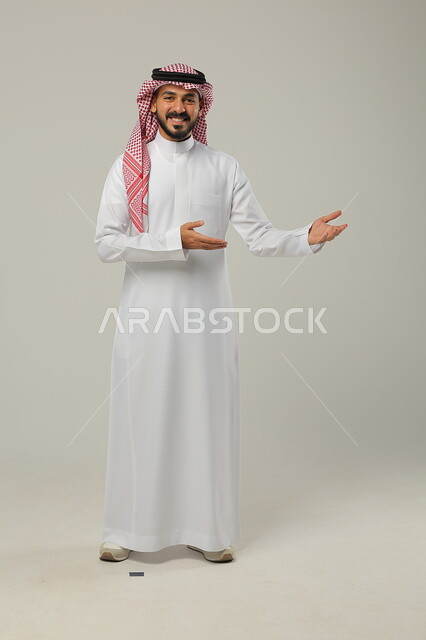 Looking at the camera with gestures of happiness and pleasure, pointing to a place, paying attention to the external appearance, portrait of a smiling Saudi Gulf Arab man wearing the traditional thobe and shemagh, full-length body portrait, gray background
