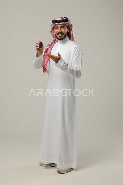 تعابير الفرح لامتلاك سيارة جديدة, النظر إلى الكاميرا بإيماءات السعادة و السرور, بورتريه لرجل عربي خليجي سعودي مبتسم يرتدي الثوب التقليدي و الشماغ يشير بيده لمفتاح التحكم عن بعد, صورة للجسم بالطول الكامل, خلفية رمادية