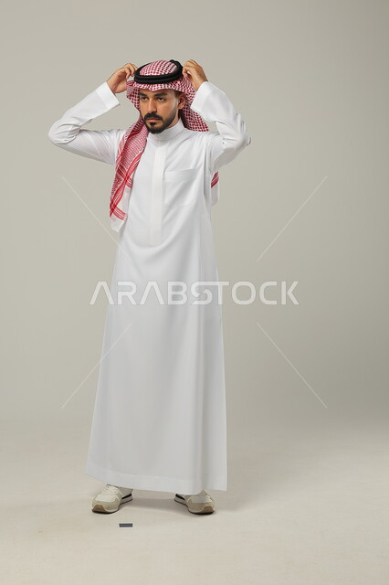 The concept of elegance and attention to external appearance, gestures of self-confidence, portrait of a Saudi Gulf Arab man wearing a traditional thobe, placing his hands on his head and adjusting the shemagh, full-length body portrait, gray background