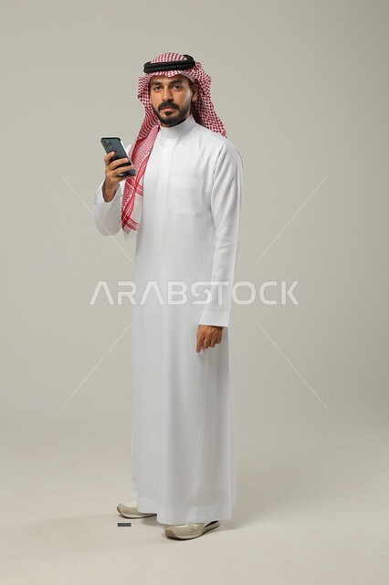 التسلية و الترفيه باستخدام الأجهزة التقنية الحديثة, التواصل مع الأهل و الأصدقاء, بورتريه لرجل عربي خليجي سعودي يرتدي الثوب التقليدي و الشماغ يمسك الهاتف المحمول بيده و ينظر إلى الكاميرا, صورة للجسم بالطول الكامل, خلفية رمادية