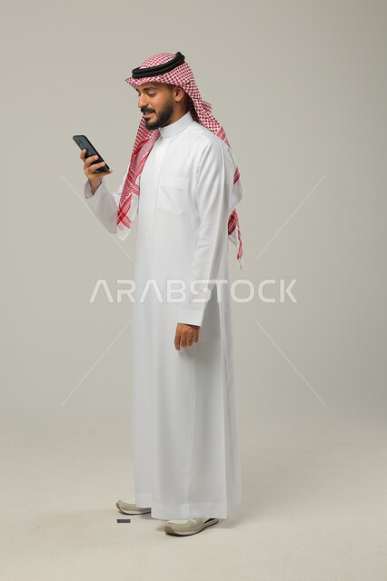 Using modern technical devices for fun and entertainment, browsing social media via mobile phone, portrait of a smiling Saudi Gulf Arab man wearing a traditional thobe and shemagh holding a mobile phone in his hand, full-length body photo, gray background