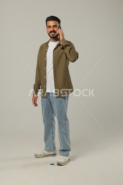 النظر إلى الكاميرا بإيماءات البشاشة، التواصل مع الأهل والأصدقاء, متابعة آخر الأخبار والمستجدات حول العالم, دمج التقنيات الحديثة والتكنولوجيا بالحياة العملية, بورتريه لشاب وسيم عربي خليجي سعودي يرتدي الزي الكاجوال يجري مكالمة هاتفية، خلفية رمادية