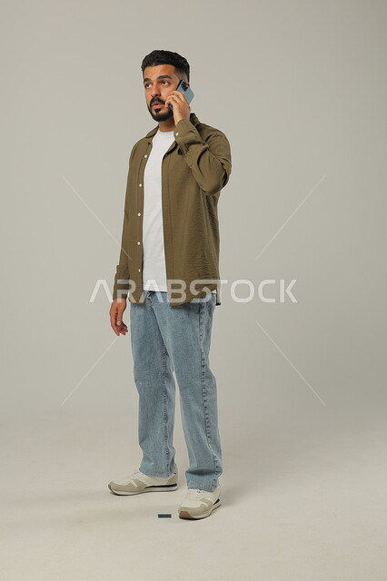 Hearing unpleasant news with expressions of annoyance, communicating with family and friends, following the latest developments around the world, integrating modern technologies and technology into practical life, portrait of a handsome Saudi Gulf Arab young man wearing casual clothes making a phone call, gray background