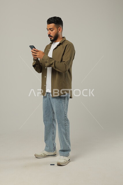 Looking at the mobile phone with integration gestures, communicating with family and friends, following the latest developments around the world, integrating modern technologies and technology into practical life, portrait of a handsome Saudi Gulf Arab young man wearing a casual outfit holding a mobile phone in his hand, body full length, gray background