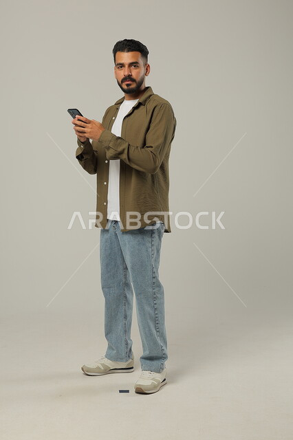 Communicating and chatting with family and friends, following the latest developments around the world, integrating modern technologies and technology into practical life, portrait of a handsome Saudi Gulf Arab young man wearing a casual outfit holding a mobile phone in his hand and looking at the camera seriously, full-length body, gray background