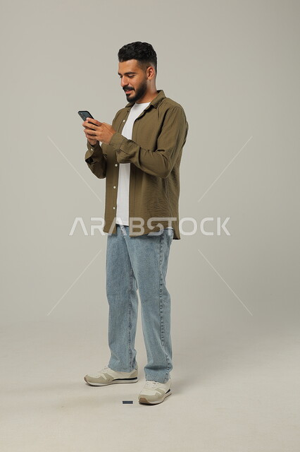 تصفح مواقع التواصل الاجتماعي، الدردشة مع الأهل والأصدقاء, متابعة آخر المستجدات حول العالم, دمج التقنيات الحديثة والتكنولوجيا بالحياة العملية, بورتريه لشاب وسيم عربي خليجي سعودي يرتدي الزي الكاجوال يمسك الهاتف المحمول بيده، الجسم بالطول الكامل، خلفية رمادية