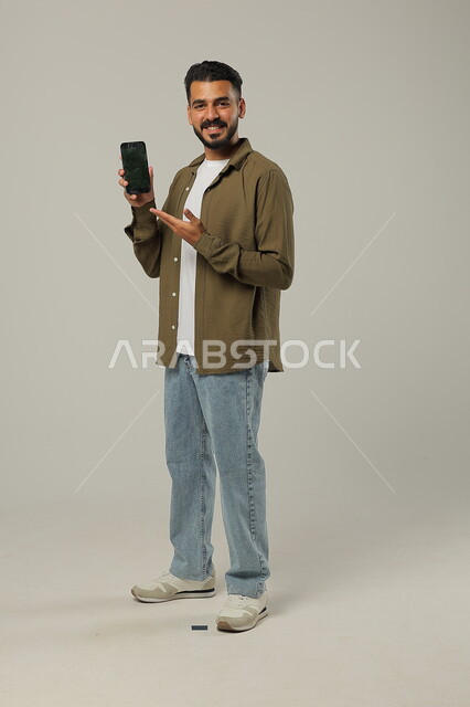الإمساك بالهاتف المحمول والإشارة للشاشة السوداء الفارغة، النظر إلى الكاميرا بإيماءات السعادة، دمج التقنيات الحديثة والتكنولوجيا بالحياة العملية, بورتريه لشاب وسيم عربي خليجي سعودي يرتدي الزي الكاجوال، الجسم بالطول الكامل، خلفية رمادية