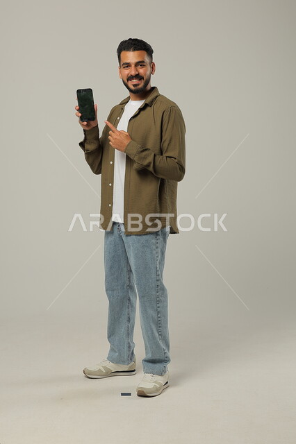 Standing and pointing at the empty black mobile screen, looking at the camera with happy gestures, integrating modern technologies and technology into practical life, portrait of a handsome Saudi Gulf Arab young man wearing a casual outfit holding a mobile phone in his hand, body full length, gray background