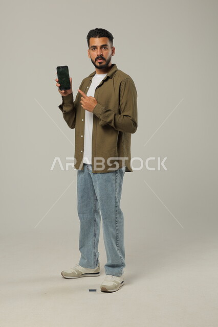 Pointing to the empty black mobile screen, looking at the camera with happy gestures, integrating modern technologies and technology into practical life, portrait of a handsome Saudi Gulf Arab young man wearing casual clothes, standing and holding a mobile phone, full-length body, gray background