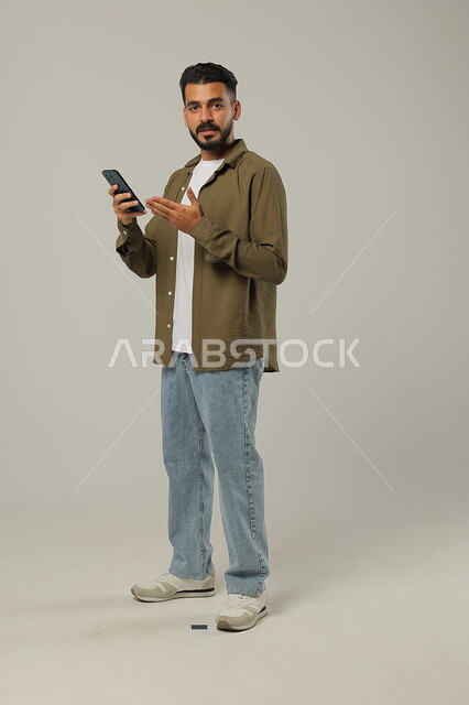 الإشارة للجوال والنظر إلى الكاميرا بإيماءات البشاشة، التواصل مع الأهل والأصدقاء, دمج التقنيات الحديثة والتكنولوجيا بالحياة العملية, بورتريه لشاب وسيم عربي خليجي سعودي يرتدي الزي الكاجوال يقف ويمسك الهاتف المحمول بيده، الجسم بالطول الكامل، خلفية رمادية