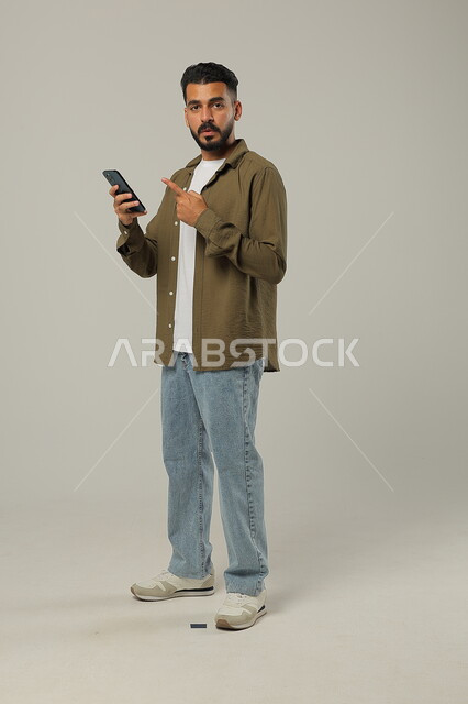 الوقوف والنظر للكاميرا بإيماءات البشاشة، التواصل مع الأهل والأصدقاء, دمج التقنيات الحديثة والتكنولوجيا بالحياة العملية, بورتريه لشاب وسيم عربي خليجي سعودي يرتدي الزي الكاجوال يمسك الهاتف المحمول بيده ويشير إليه، الجسم بالطول الكامل، خلفية رمادية