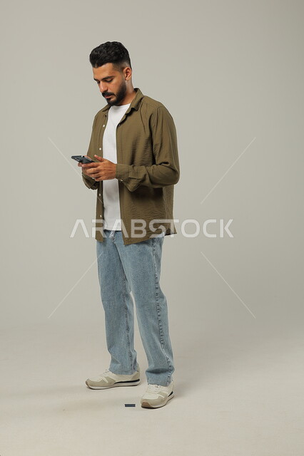  إيماءات الاندماج مع الجوال، التواصل مع الأهل والأصدقاء, متابعة آخر المستجدات حول العالم, دمج التقنيات الحديثة والتكنولوجيا بالحياة العملية, بورتريه لشاب وسيم عربي خليجي سعودي يرتدي الزي الكاجوال يمسك الهاتف المحمول بيده، الجسم بالطول الكامل، خلفية رمادية