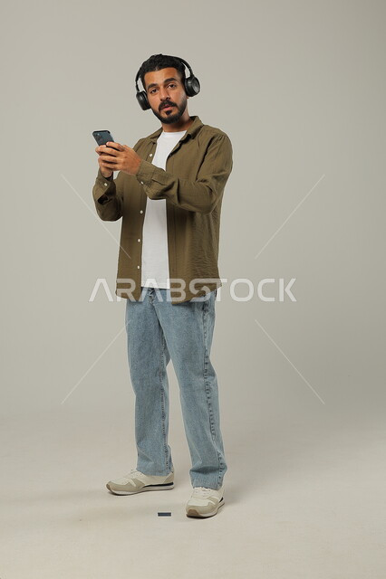 النظر للكاميرا عند الاستماع إلى الموسيقا والتسجيلات الصوتية، الاستفادة من الجوال والأجهزة التكنولوجية، شاب عربي خليجي سعودي يرتدي الزي الكاجوال ويضع سماعة الرأس بإيماءات الاندماج, استخدام تقنية الهيدفون، جسم بالطول الكامل, خلفية رمادية