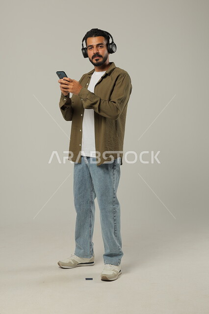 الاستمتاع بتقنية الهيدفون، استخدام الجوال والأجهزة التكنولوجية، إيماءات الاندماج والتركيز عند الاستماع إلى الموسيقا والمقاطع الصوتية، شاب عربي خليجي سعودي يرتدي الزي الكاجوال ويضع سماعة الرأس, جسم بالطول الكامل, خلفية رمادية
