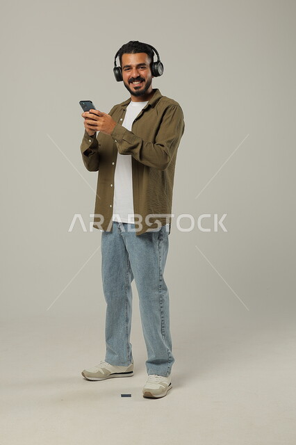 استخدام الجوال والأجهزة التكنولوجية، إيماءات الاندماج والتركيز عند الاستماع إلى الموسيقا والمقاطع الصوتية، شاب عربي خليجي سعودي يرتدي الزي الكاجوال ويضع سماعة الرأس بسعادة, الاستمتاع بتقنية الهيدفون، جسم بالطول الكامل, خلفية رمادية
