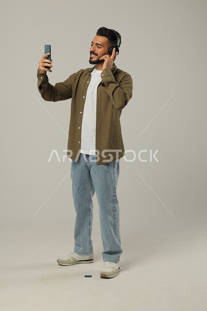 استخدام تقنية الهيدفون، الاستفادة من الجوال والأجهزة التكنولوجية، الاستماع إلى الموسيقا والتسجيلات الصوتية، شاب عربي خليجي سعودي يرتدي الزي الكاجوال ويضع سماعة الرأس بإيماءات السعادة, جسم بالطول الكامل, خلفية رمادية