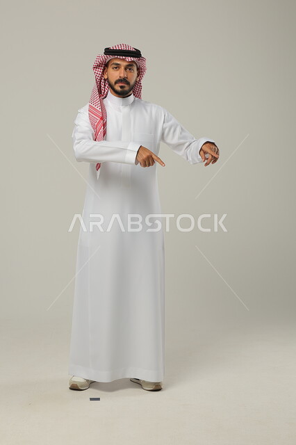 الوقوف باستقامة و اعتدال, الإشارة إلى شيء ما بالأسفل, بورتريه لرجل عربي خليجي سعودي يرتدي الثوب التقليدي و الشماغ يشير بإصبعي السبابة و ينظر إلى الكاميرا, صورة للجسم بالطول الكامل, خلفية رمادية