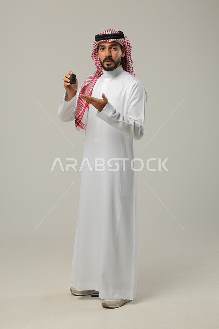 النظر إلى الكاميرا بتعابير الدهشة, الفرح لامتلاك سيارة جديدة, بورتريه لرجل عربي خليجي سعودي يرتدي الثوب التقليدي و الشماغ يمسك بيده مفتاح التحكم عن بعد و يشير اليه, صورة للجسم بالطول الكامل, خلفية رمادية