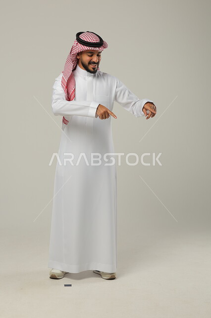 النظر بإيماءات السعادة و السرور, الإشارة إلى شيء ما بالأسفل, بورتريه لرجل عربي خليجي سعودي مبتسم يرتدي الثوب التقليدي و الشماغ يشير بإصبعي السبابة, صورة للجسم بالطول الكامل, خلفية رمادية