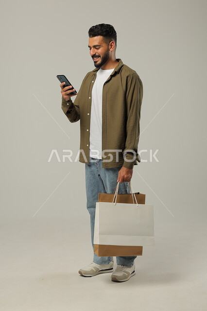 عمليات الشراء الالكتروني عبر الانترنت, مفهوم التسوق عن بعد, بورتريه لشاب عربي خليجي سعودي يرتدي الزي الكاجوال يمسك بيده أكياس ورقية ملونة, الإمساك بالهاتف المحمول وإتمام عملية الطلبات, التبضع والاستفادة من العروض والتخفيضات, جسم بالطول الكامل, خلفية رمادية