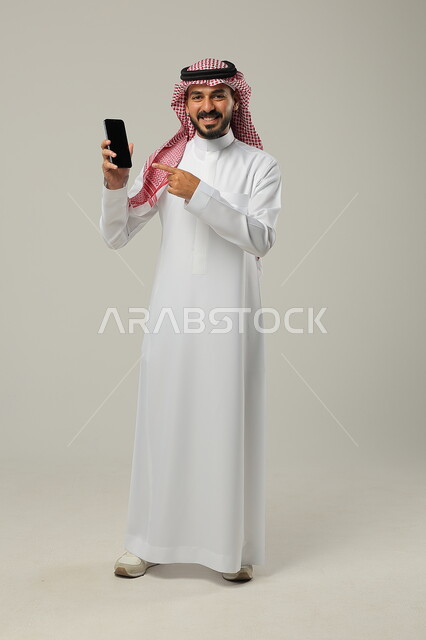 الإشارة بإصبع السبابة إلى الجوال, استخدام الأجهزة التقنية الحديثة و المتطورة, بورتريه لرجل عربي خليجي سعودي مبتسم يرتدي الثوب التقليدي و الشماغ يمسك الهاتف المحمول بيده بشاشة سوداء فارغة, صورة للجسم بالطول الكامل, خلفية رمادية