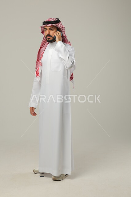إيماءات الاستياء بسماع خبر ما, إجراء مكالمة هاتفية عبر الجوال, بورتريه لرجل عربي خليجي سعودي يرتدي الثوب التقليدي و الشماغ يمسك الهاتف المحمول بيده, صورة للجسم بالطول الكامل, خلفية رمادية