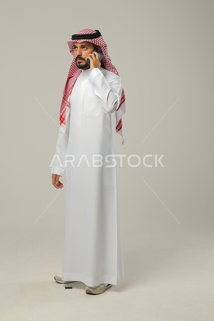 Looking up with an expression of dissatisfaction, making a phone call on a mobile phone, portrait of a Saudi Gulf Arab man wearing a traditional thobe and shemagh holding a mobile phone in his hand, full-length body portrait, gray background
