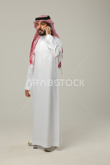 إجراء مكالمة هاتفية عبر الجوال, التواصل و التحدث مع العائلة و الأصدقاء, بورتريه لرجل عربي خليجي سعودي يرتدي الثوب التقليدي و الشماغ يمسك الهاتف المحمول بيده, صورة للجسم بالطول الكامل, خلفية رمادية