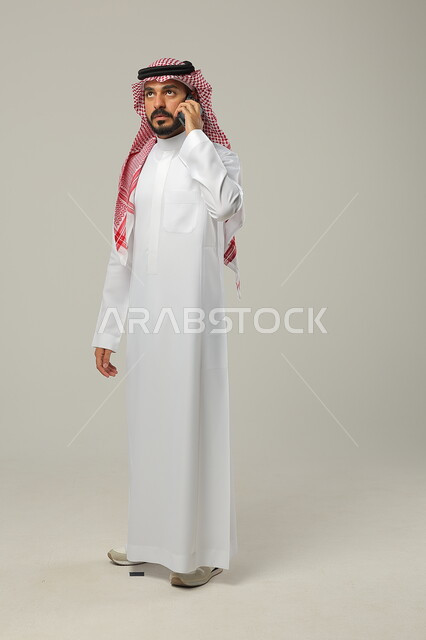 تبادل الأحاديث و الأخبار مع العائلة و الأصدقاء, إجراء اتصال هاتفي عبر الجوال, بورتريه لرجل عربي خليجي سعودي يرتدي الثوب التقليدي و الشماغ يمسك الهاتف المحمول بيده و ينظر إلى الأعلى, صورة للجسم بالطول الكامل, خلفية رمادية