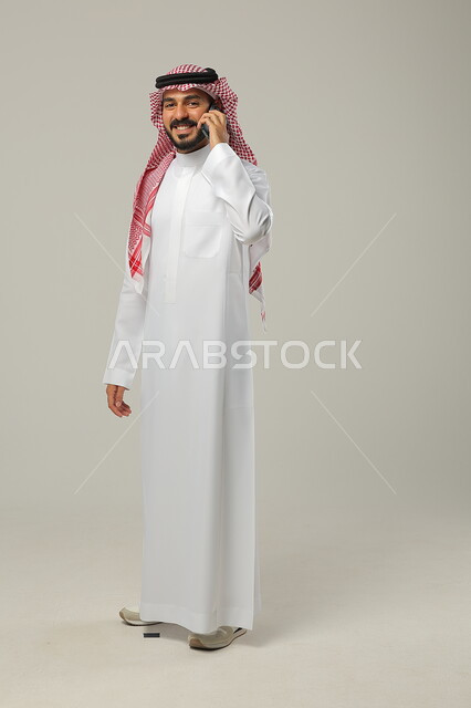 تعابير الفرح بالتواصل مع العائلة و الأصدقاء, إجراء اتصال هاتفي عبر الجوال, تبادل الأحاديث و الأخبار الجيدة, بورتريه لرجل عربي خليجي سعودي مبتسم يرتدي الثوب التقليدي و الشماغ يمسك الهاتف المحمول بيده, صورة للجسم بالطول الكامل, خلفية رمادية
