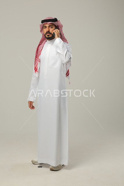 إجراء اتصال هاتفي عبر الجوال, التواصل مع العائلة و الأصدقاء, تبادل الأحاديث و الأخبار الجيدة, بورتريه لرجل عربي خليجي سعودي يرتدي الثوب التقليدي و الشماغ يمسك الهاتف المحمول بيده, صورة للجسم بالطول الكامل, خلفية رمادية