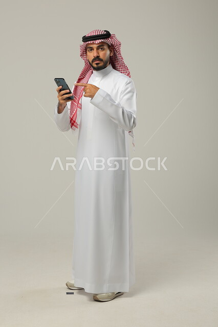 Chat and communicate with friends, pointing with the index finger at the mobile phone, portrait of a Saudi Gulf Arab man wearing the traditional dress and shemagh, holding the mobile phone in his hand and looking at the camera, full-length body photo, gray background