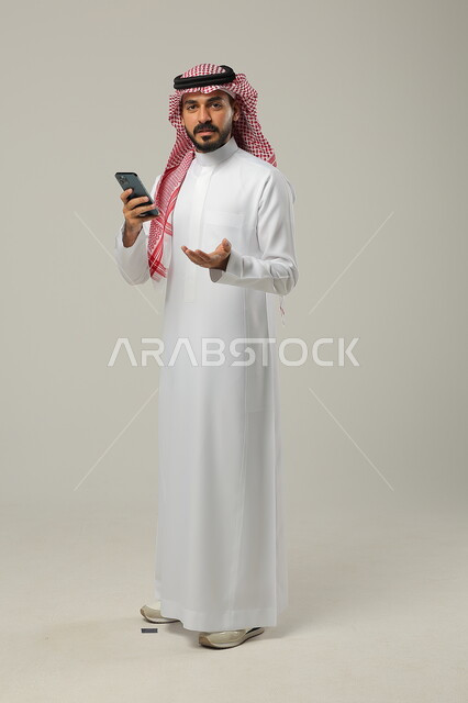 Using modern and advanced technology, looking at the camera with surprised and questioning gestures, portrait of a Saudi Gulf Arab man wearing a traditional thobe and shemagh, holding a mobile phone in his hand, full-length body portrait, gray background