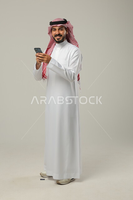 Looking at the camera with gestures of happiness and pleasure, using modern and advanced technology, portrait of a smiling Saudi Gulf Arab man wearing a traditional thobe and shemagh holding a mobile phone in his hand, full-length body portrait, gray background