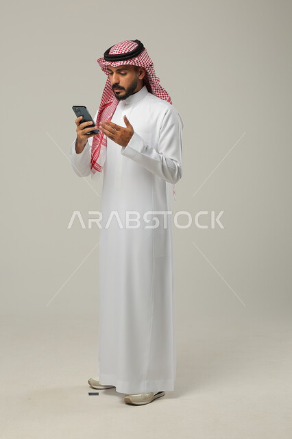 التواصل مع العائلة و الأصدقاء عبر الجوال, استخدام الأجهزة التقنية المتطورة, بورتريه لرجل عربي خليجي سعودي يرتدي الثوب التقليدي و الشماغ يمسك الهاتف المحمول بيده وينظر اليه, صورة للجسم بالطول الكامل, خلفية رمادية