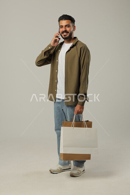  تأكيد الطلبات أونلاين بإجراء مكالمات هاتفية، استخدام الجوال للتسوق عن بعد, الشراء الالكتروني عبر الانترنت, بورتريه لشاب عربي خليجي سعودي يرتدي الزي الكاجوال يمسك بيده أكياس ورقية ملونة, التبضع والاستفادة من العروض والتخفيضات, جسم بالطول الكامل, خلفية رمادية