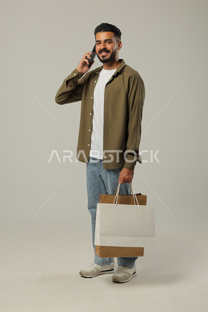  إجراء مكالمة هاتفية لتأكيد الطلب، استخدام الهاتف المحمول للتسوق عن بعد, الشراء الالكتروني عبر الانترنت, بورتريه لشاب عربي خليجي سعودي يرتدي الزي الكاجوال يمسك بيده أكياس ورقية ملونة, التبضع والاستفادة من العروض والتخفيضات, جسم بالطول الكامل, خلفية رمادية
