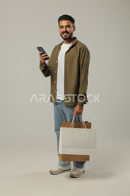 Completing the ordering process using a mobile phone, the concept of remote shopping, electronic purchases via the Internet, portrait of a Saudi Gulf Arab young man wearing a casual outfit holding colored paper bags in his hand, shopping and taking advantage of offers and discounts, full-length body, gray background