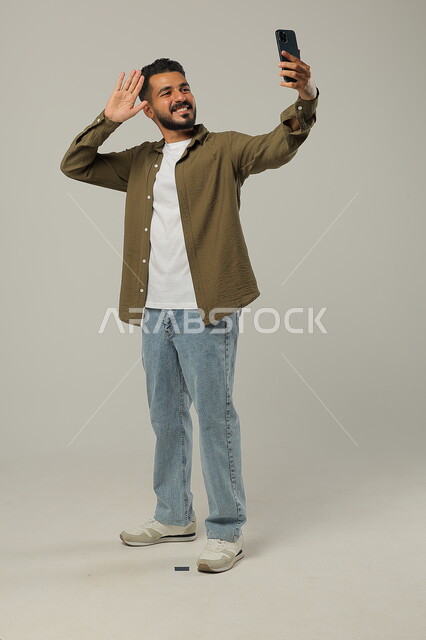 التقاط صور السيلفي التذكارية, الأناقة والاهتمام بالمظهر الخارجي, بورتريه لشاب وسيم عربي خليجي سعودي يرتدي الزي الكاجوال يقف ويمسك الهاتف المحمول بيده بإيماءات السعادة، الجسم بالطول الكامل، خلفية رمادية