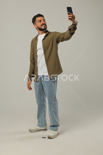 الأناقة والاهتمام بالمظهر الخارجي, التقاط صور السيلفي التذكارية, بورتريه لشاب وسيم عربي خليجي سعودي يرتدي الزي الكاجوال يقف ويمسك الهاتف المحمول بيده بإيماءات السعادة، الجسم بالطول الكامل، خلفية رمادية