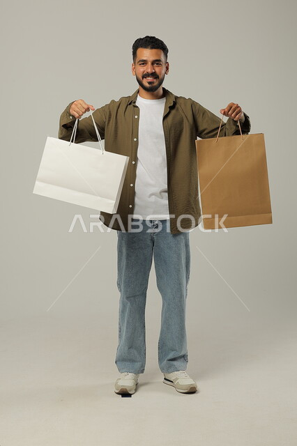 الحصول على الحسومات والتنزيلات, مواسم التخفيضات في السعودية, بورتريه لشاب عربي خليجي سعودي يرتدي الزي الكاجوال يمسك بيده أكياس ورقية, المشتريات التجارية والهدايا, التبضع وأخذ الحاجات اللازمة والضرورية, متعة التسوق في المملكة, جسم بالطول الكامل, خلفية رمادية