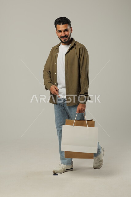 عرض أكياس الهدايا والإشارة إلى الاستفادة من الحسومات والتصفيات, بورتريه لشاب عربي خليجي سعودي يرتدي الزي الكاجوال يمسك بيده حقائب ورقية تجارية بإيماءات السعادة, التبضع وأخذ الحاجات الضرورية, متعة التسوق في المملكة, جسم بالطول الكامل, خلفية رمادية