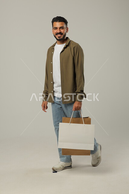 التبضع وأخذ الحاجات اللازمة والضرورية, المشتريات التجارية والهدايا, مواسم التخفيضات في السعودية, الاستفادة من الحسومات والتصفيات, بورتريه لشاب عربي خليجي سعودي يرتدي الزي الكاجوال يمسك بيده أكياس ورقية, متعة التسوق في المملكة, جسم بالطول الكامل, خلفية رمادية