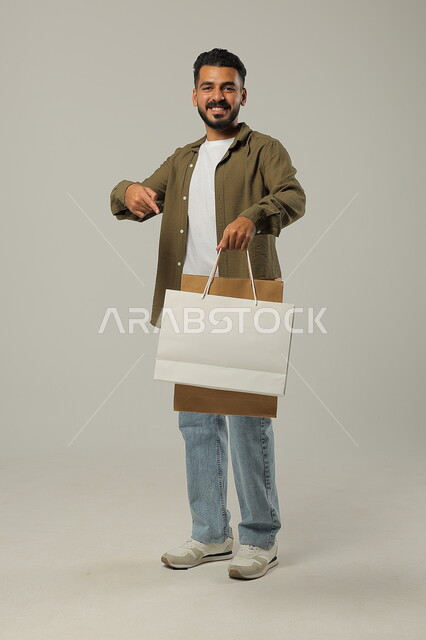 النظر للكاميرا والإشارة إلى حقائب المشتريات التجارية بسعادة, الاستفادة من الحسومات والتصفيات, بورتريه لشاب عربي خليجي سعودي يرتدي الزي الكاجوال يمسك بيده أكياس ورقية, التبضع وأخذ الحاجات الضرورية, متعة التسوق في المملكة, جسم بالطول الكامل, خلفية رمادية