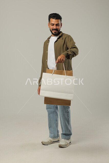 Sales seasons in Saudi Arabia, taking advantage of discounts and clearances, a portrait of a Saudi Gulf Arab young man wearing a casual outfit holding paper bags in his hand, commercial purchases and gifts, shopping and taking necessary and necessary things, the joy of shopping in the Kingdom, full-length body, gray background