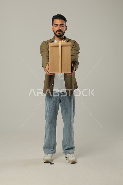 Celebrating holidays and happy occasions, giving a gift with enthusiasm and happiness, giving gifts to family and close friends, portrait of a handsome Saudi Gulf Arab young man wearing a casual outfit holding a wrapped gift box in his hands, full-length body, gray background
